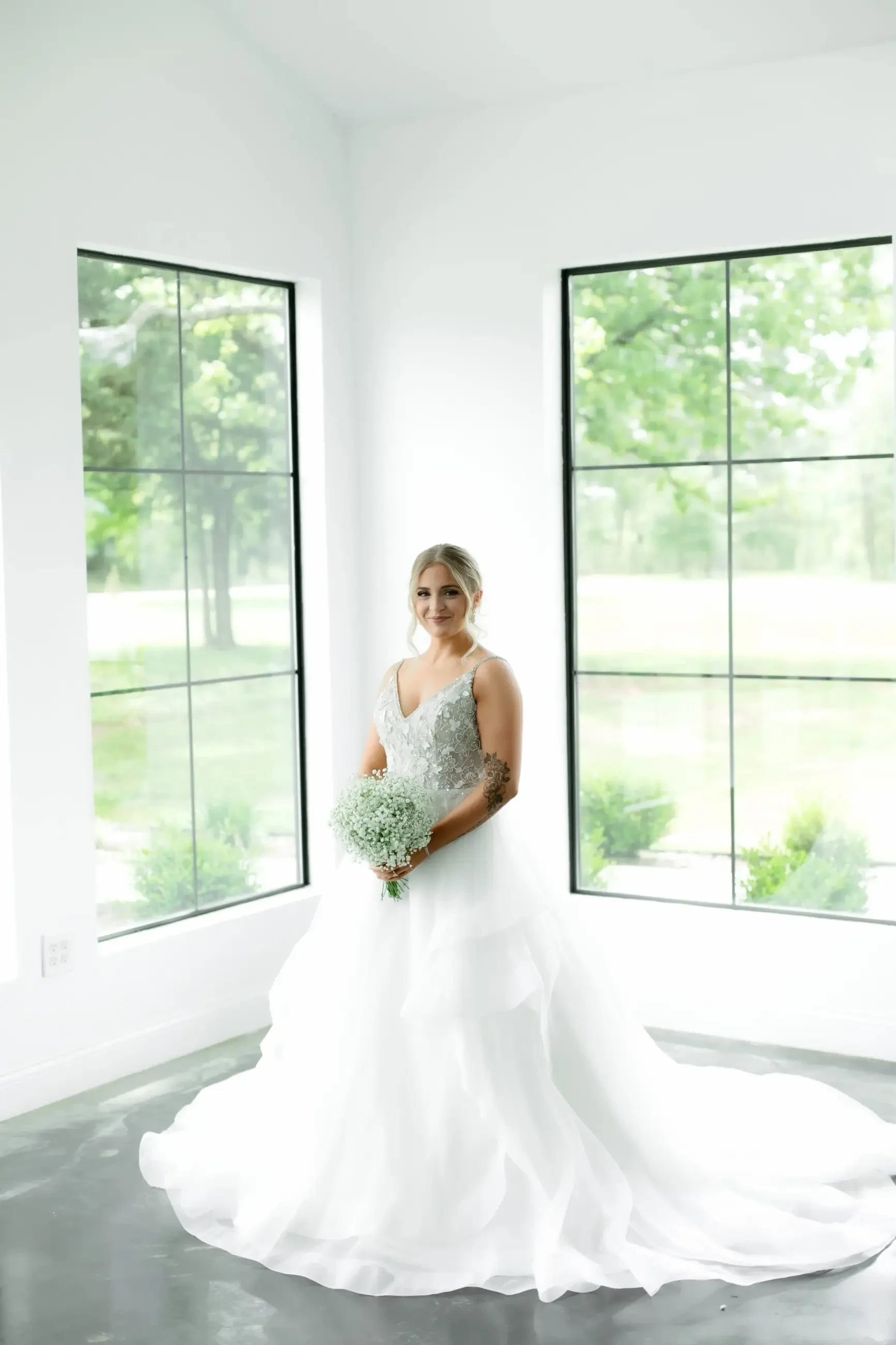 A bride in a white gown holds a bouquet, standing in a bright room with large windows. The room is filled with natural light and greenery outside creates a serene backdrop.