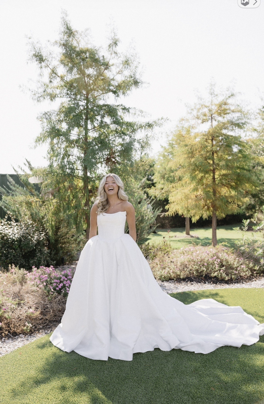 A joyful bride in a strapless white gown stands on lush green grass, surrounded by trees and flowers. The sunny day highlights her radiant smile.